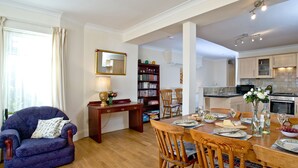 A dining area with a wooden table set for four, surrounded by chairs, a bookshelf, and a window with curtains. - A spacious apartment overlooking Goodrington Park with sea views. (Paignton)