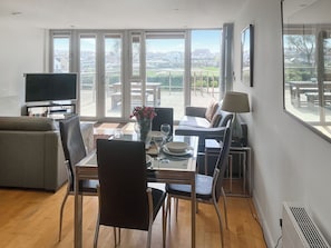 A modern living room with a glass dining table, chairs, a sofa, a TV, and large windows offering a view of the outdoors.