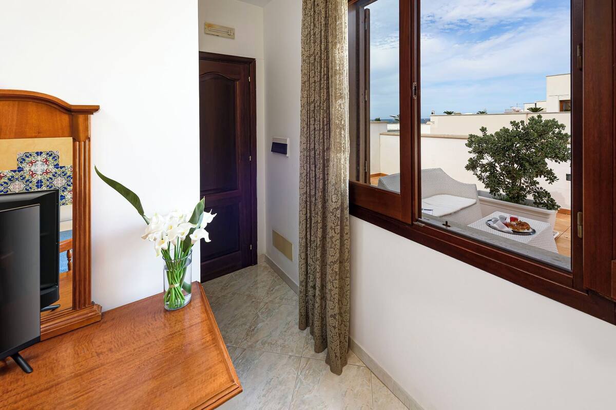 A hotel room with a wooden desk, a vase of flowers, a window with a view of the city, and a door leading to another room.