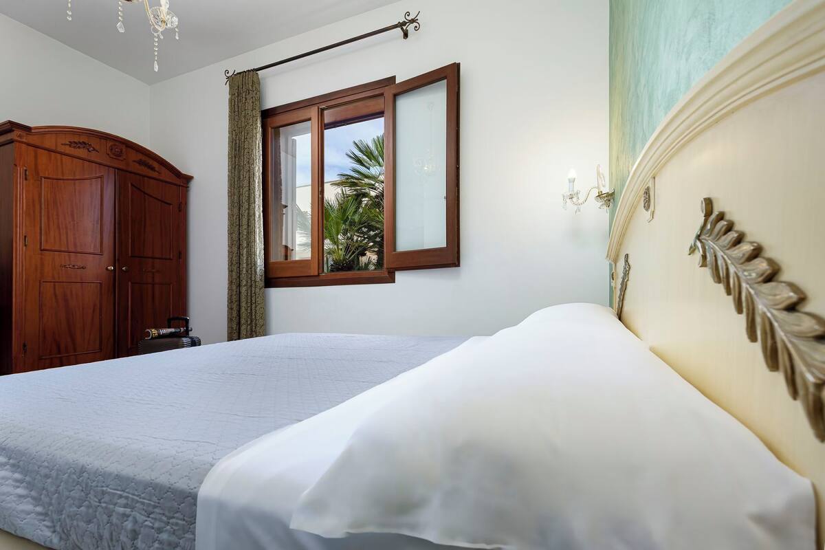 A bedroom with a bed, a wooden wardrobe, a window with a view of palm trees, and a chandelier.