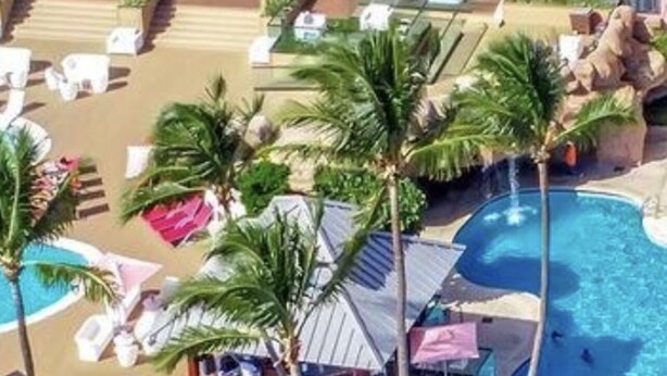 Un complexe hôtelier doté d’une piscine, de chaises longues, de palmiers et d’un espace abrité avec des parasols.