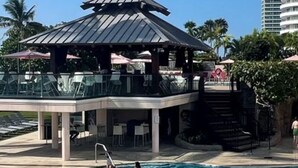Une piscine avec des personnes dans l’eau, un espace couvert avec un toit, et des palmiers en arrière-plan.