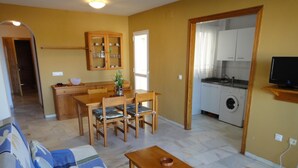 A living room with a sofa, wooden dining table, and chairs. There is a kitchen area with a washing machine and cabinets.