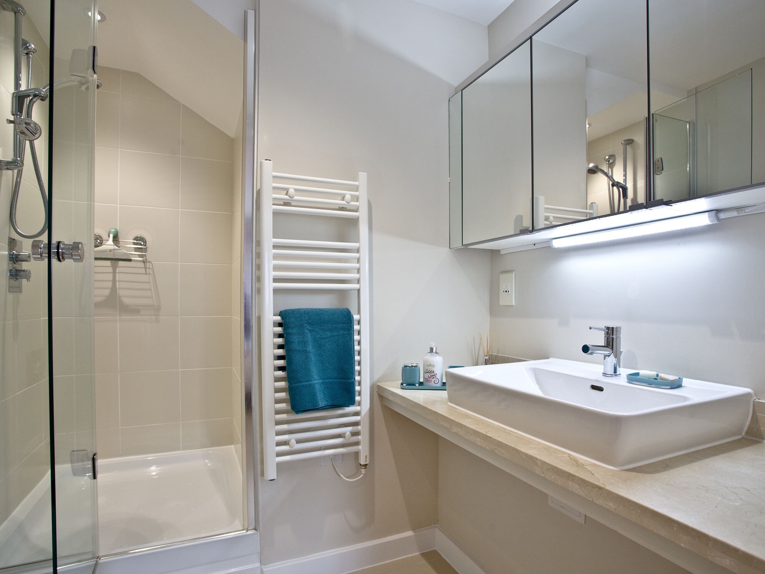 Un baño moderno con mampara de vidrio, un lavamanos blanco y un perchero con una toalla azul.