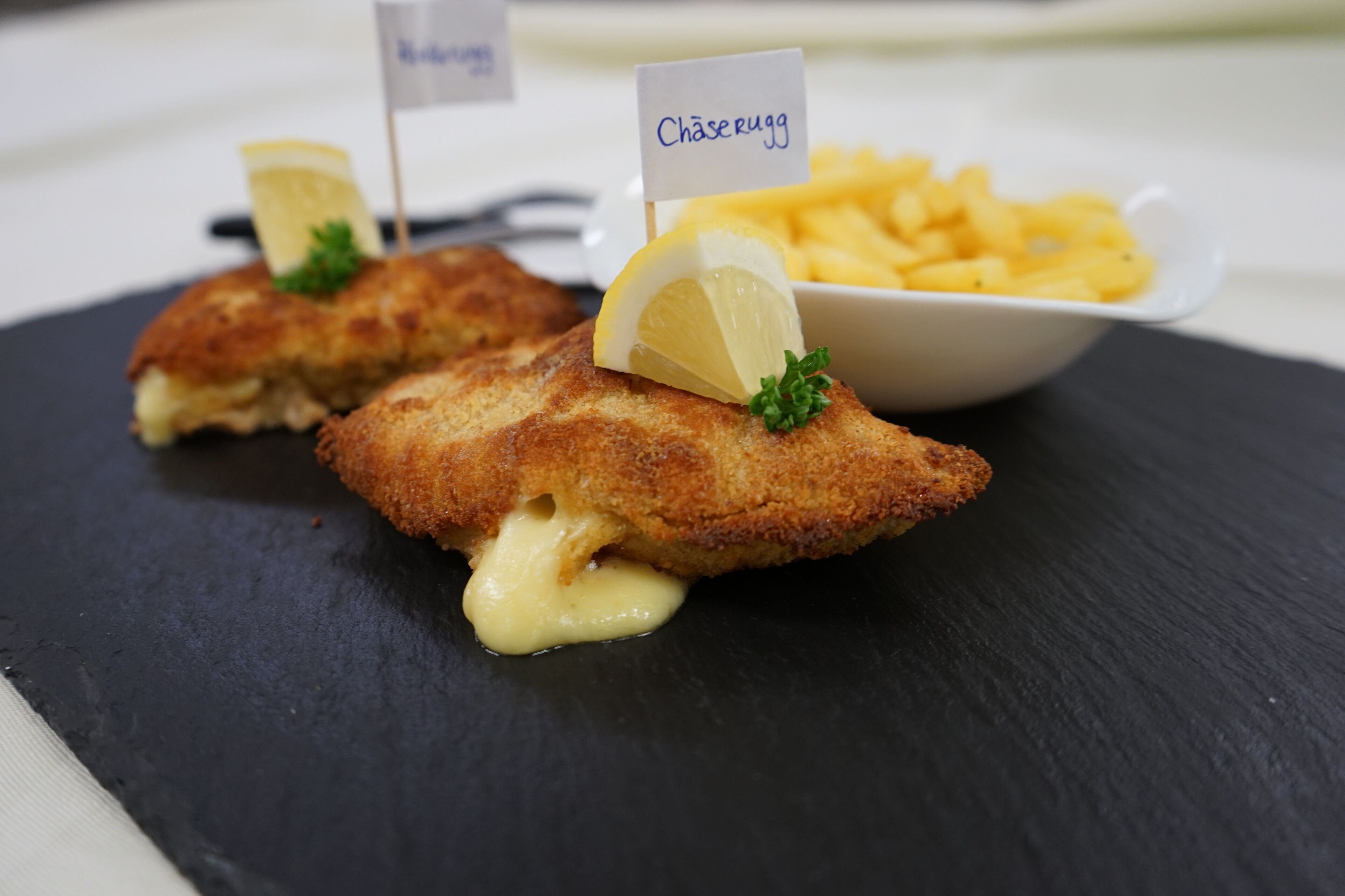 Dos filetes de pescado rebozados, con adorno de limón y una pequeña bandera, servidos sobre un plato de pizarra negra, acompañados de una cesta de papas fritas.