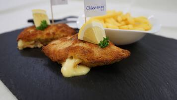 Dos filetes de pescado rebozados, con adorno de limón y una pequeña bandera, servidos sobre un plato de pizarra negra, acompañados de una cesta de papas fritas.