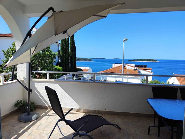 A balcony with a chair and table, overlooking a coastal view with buildings and trees.
