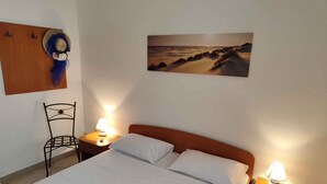 A neatly made bed with white linens, two bedside tables with lamps, a metal chair, and a painting of a beach scene on the wall. - Holiday apartment with a view of the lake (Primošten)