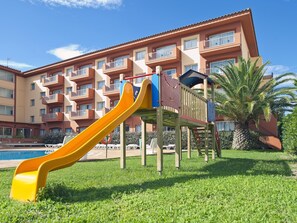 Un terrain de jeux avec un toboggan jaune et des structures d’escalade, situé devant un immeuble à plusieurs étages doté de balcons, avec une piscine en arrière-plan.