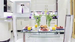 A modern kitchen with white cabinets, a glass dining table, and a breakfast setup including fruits, cookies, and juice. - 2 bedroom accommodation in Torquay (Torquay)