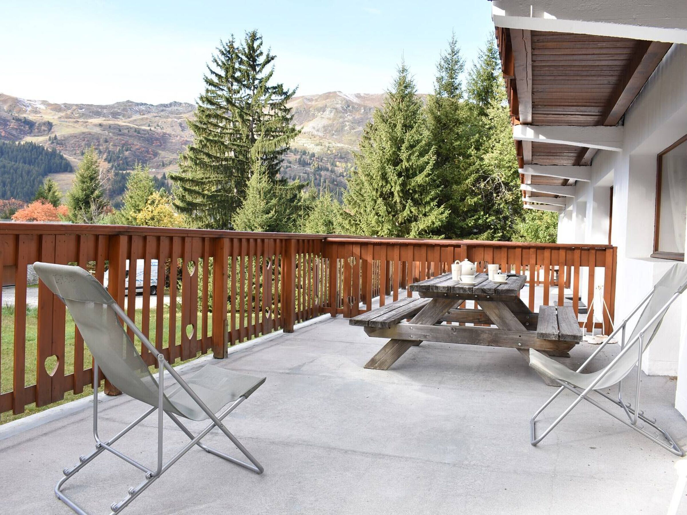 A balcony with a wooden fence, two folding chairs, a picnic table, and a view of mountains and trees.