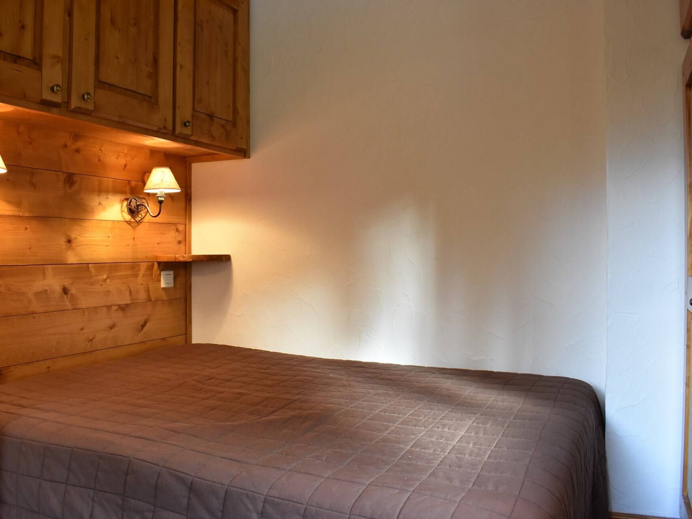 Une chambre avec une tête de lit en bois, un lit recouvert d’une courtepointe marron et une lampe fixée au mur.
