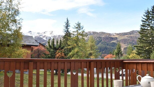 Eine Holzterrasse mit einem für Tee gedecktem Tisch, mit Blick auf eine gebirgige Landschaft mit Herbstbäumen und eine Hütte.