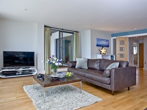 A modern living room with a leather sofa, a flat-screen TV, a coffee table with a vase of flowers, and a large window with curtains. - The Penthouse Bay View (Westward Ho!)