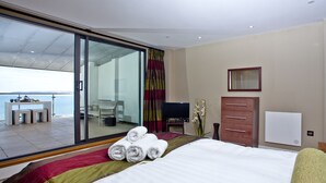 A hotel room with a bed, a TV, a dresser, and a view of the sea through a large window. - The Penthouse Bay View (Westward Ho!)