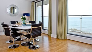 A modern room with a dining table, chairs, a mirror, and large windows with curtains. - The Penthouse Bay View (Westward Ho!)