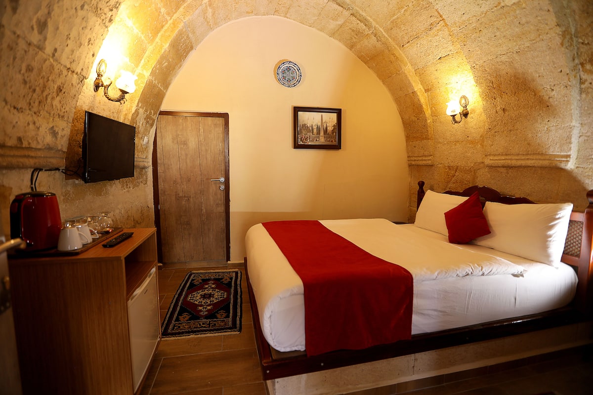 A hotel room with a wooden bed, a red throw, a flat-screen TV, and a wooden desk with a kettle and cups.