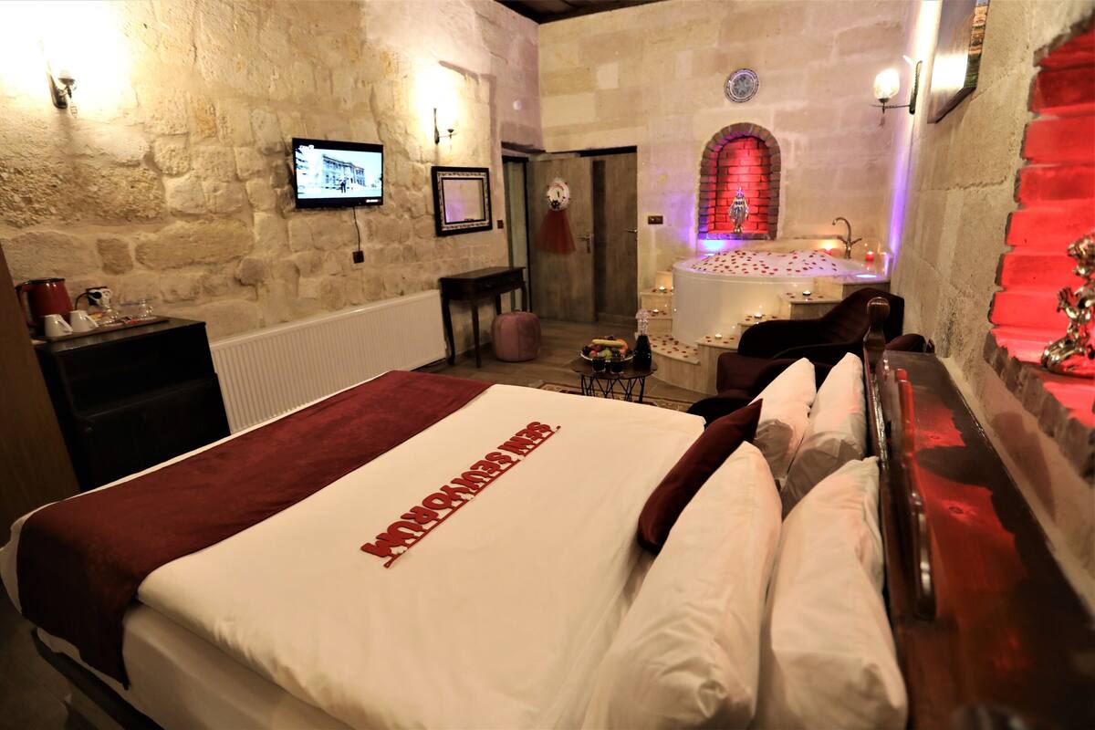 A hotel room with a stone wall, a bed with a red and white bedspread, a television, a small table with a fruit bowl, and a bathroom with a red-lit archway.