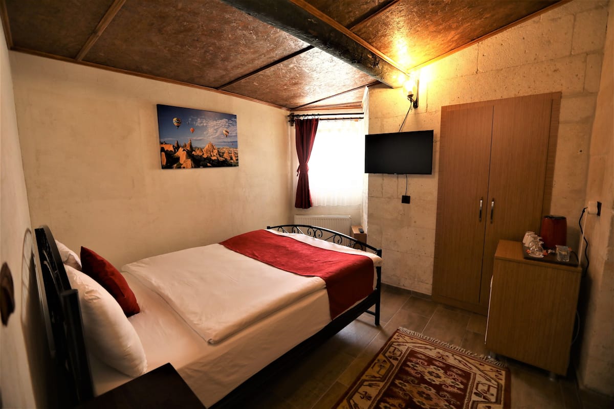 A bedroom with a bed, a TV mounted on the wall, a wooden wardrobe, and a window with curtains.