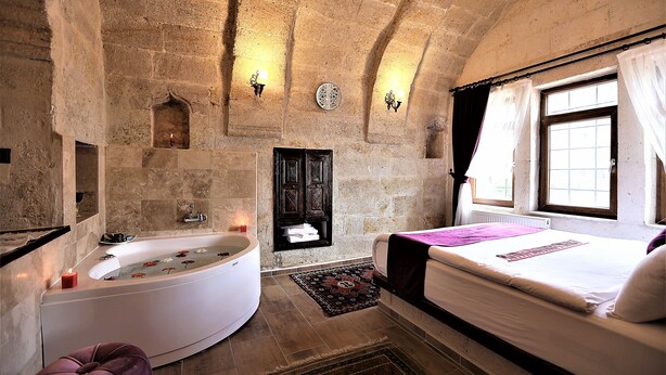 A stone-walled room with a large bathtub, a bed, and a small window with curtains. - Orion Cave Hotel (Ürgüp)