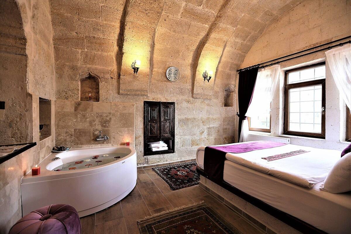 A stone-walled room with a bathtub, a bed, and a window with curtains.
