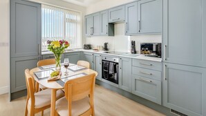 A modern kitchen with grey cabinets, a dining table with chairs, and a window with blinds. - Ideally positioned apartment with large balcony overlooking Goodrington Beach. (Paignton)