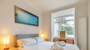 A neatly made bed with white linens, two bedside lamps, a wooden nightstand with drawers, a large window with blinds, and a framed painting on the wall. - Ideally positioned apartment with large balcony overlooking Goodrington Beach. (Paignton)