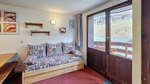 Una habitació acollidora amb una banca de fusta, un sofà estampat, una estant penjada a la paret i una finestra gran que ofereix vistes a edificis i vegetació.