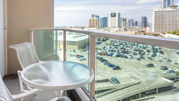 A balcony with a glass table and chairs, overlooking a parking lot and a cityscape with modern buildings. - BEST RATE!  ADA Studio Ala Moana Condominium (AH01) (Honolulu)