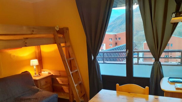 Ein Zimmer mit einem hölzernen Etagenbett, einem kleinen Tisch, einem Stuhl, einer Lampe und Blick auf Gebäude durch ein Fenster.