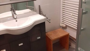 Ein Badezimmer mit einem weißen Waschbecken, dunklen Schränken, einem hölzernen Hocker und einer Duschkabine aus Glas.