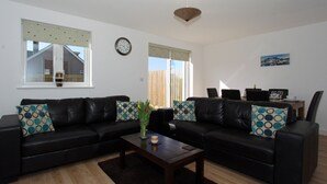 A living room with two black leather sofas, a wooden coffee table, and a dining area with a table and chairs. - 4 bedroom accommodation in Padstow (Padstow)