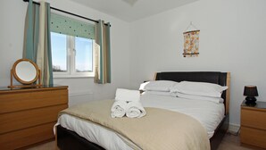 A neatly made bed with white linens and rolled towels, a wooden dresser, a bedside table with a lamp, and a window with curtains. - 4 bedroom accommodation in Padstow (Padstow)