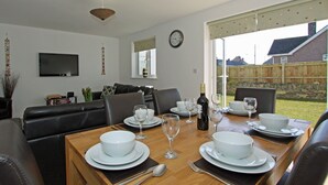 A dining area with a wooden table set for four, surrounded by leather chairs. A television is mounted on the wall, and a clock is visible. - 4 bedroom accommodation in Padstow (Padstow)