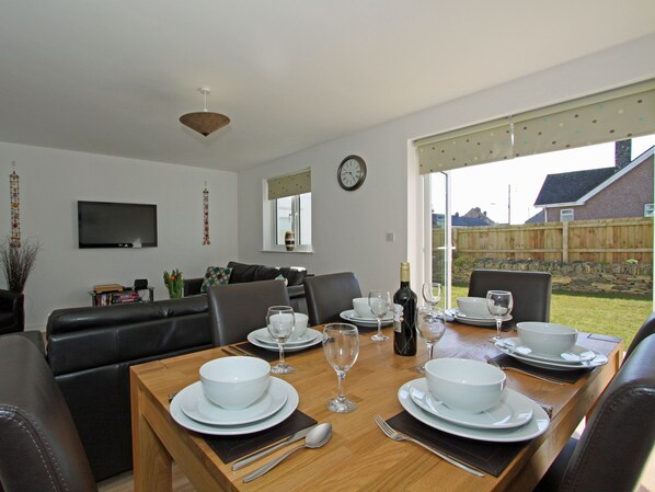 A dining area with a wooden table set for four, surrounded by leather chairs. A television is mounted on the wall, and a clock is visible.