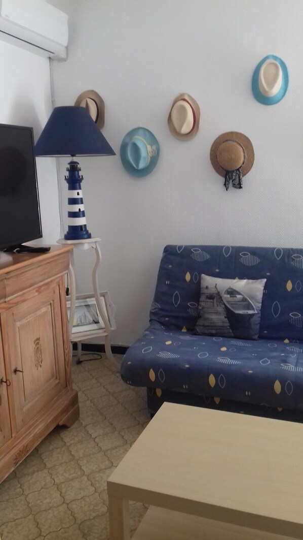 A room with a wooden cabinet, a television, a blue patterned sofa, a small table, and a wall with several hats.