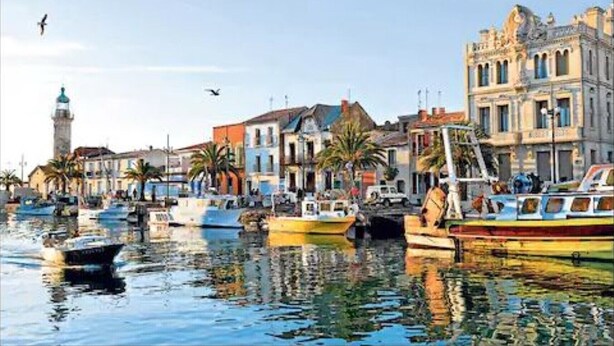 A harbor with boats docked, buildings with classical architecture, and a lighthouse in the background.
