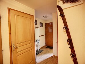 Uma porta de madeira dá para um corredor com chão de madeira, uma porta com um pequeno janelão quadrado e um suporte para esquis na parede.