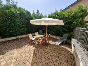 Een terras met een tafel en stoelen onder een overkapping, omringd door groen en een geplaveid terras.