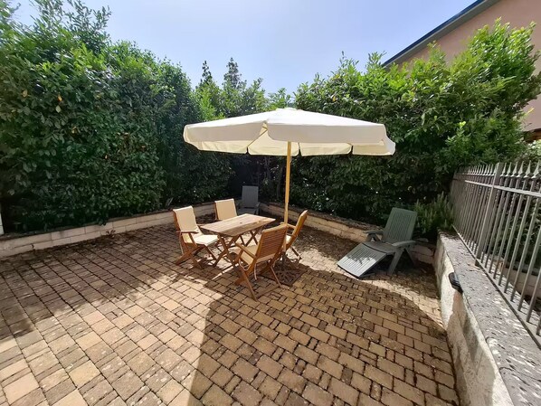 Un patio con mesa y sillas bajo un toldo, rodeado de follaje y piso de ladrillo.
