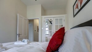 Un lit bien fait, avec du linge de lit blanc et un coussin rouge, une porte blanche donnant sur une autre pièce, et un tableau encadré accroché au mur.