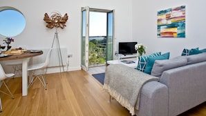 A modern living room with a grey sofa, a dining table with chairs, a TV, and a balcony with a view.