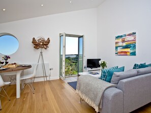 Una sala de estar moderna con un sofá gris, una mesa de comedor con sillas, un televisor y un balcón con vistas.