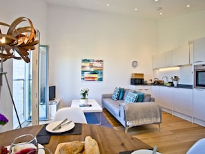 A modern apartment with a dining area, a living room with a sofa, a kitchen with white cabinets, and a large abstract painting on the wall.