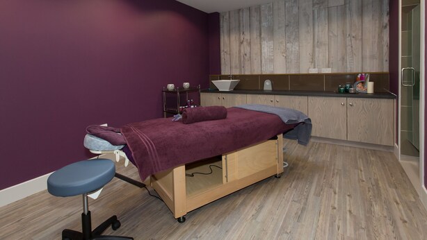 A room with a wooden floor, a bed with a purple blanket, a small table with a bowl, a mirror, and a wooden cabinet.