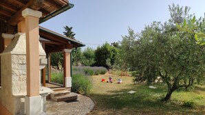 A stone-paved patio with steps leading to a covered area, surrounded by a grassy garden with trees and shrubs. - House Kras (Kras)