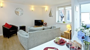 A living room with a grey sofa, a black armchair, a TV, a dining table with a plate of grapes and wine glasses, and a window with blinds.