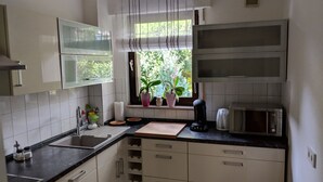 Eine kleine Küche mit weißen Schränken, einer schwarzen Arbeitsplatte, einem Fenster mit Blumentöpfen, einer Mikrowelle und einem Wasserkocher.