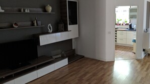 Ein modernes Wohnzimmer mit Flachbildfernseher, Holzböden und einer durch einen offenen Durchgang erkennbaren Küche.