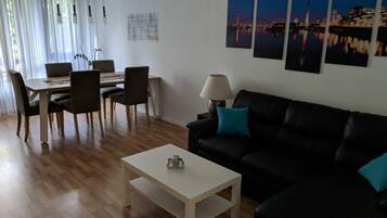 Ein modernes Wohnzimmer mit einem schwarzen Ledersofa, einem weißen Couchtisch und einem Essbereich mit Holztisch und Stühlen. An der Wand hängen vier gerahmte Bilder, und auf einem Beistelltisch steht eine Lampe.